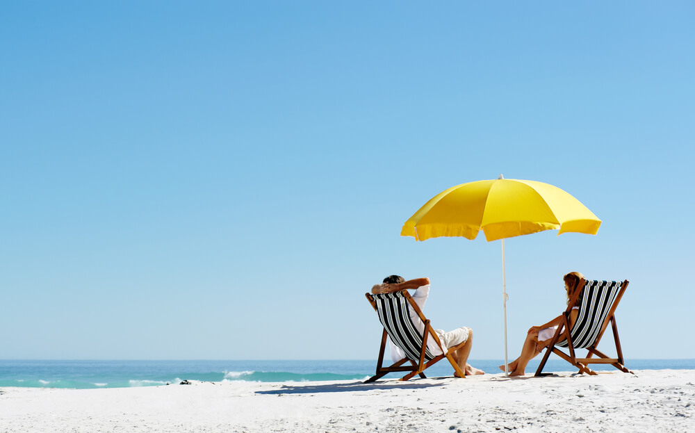 Couple sitting on beach chairs under an umbrella on the beach: living without varicose veins this summer.