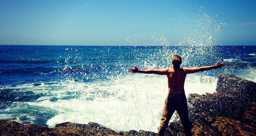 A shirtless man, with arms outstretched, getting hit by an ocean wave: spider vein symptom facts