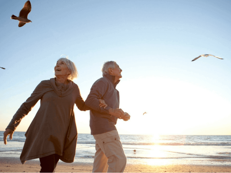 An elderly couple smiling and running on a beach, happy they got vein disease treatment.