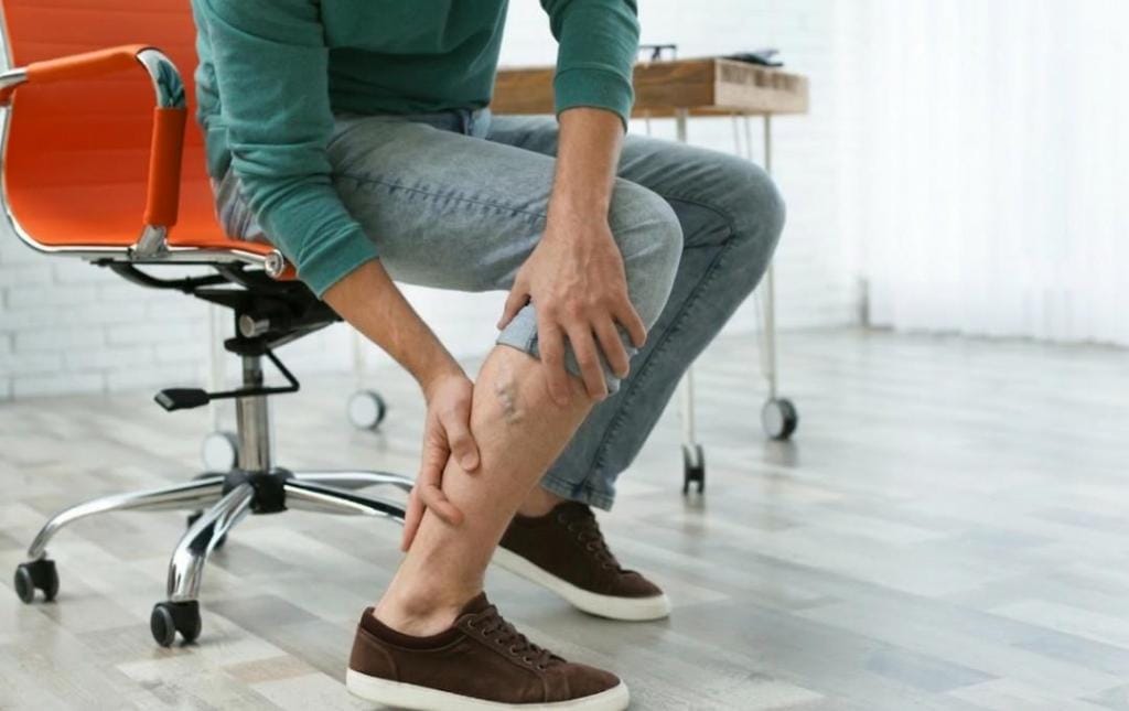 Should You Be Worried About Chronic Venous Insufficiency? Know Your Risk 7 Man sitting behind his desk and holding his leg with visible varicose veins, showing signs of chronic venous insufficiency.